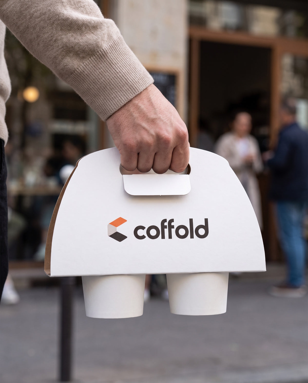 A hand carrying a Coffold two-cup drinks carrier past a cafe doorway, both takeaway cups held upright
